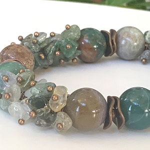 K&ouml;nnte beinhalten: Ein gr&uuml;nes, braunes und wei&szlig;es Edelstein-Armband mit Kupferdraht-Akzenten. Das Armband verf&uuml;gt &uuml;ber eine Vielzahl von Formen und Gr&ouml;&szlig;en von Edelsteinen, darunter runde, ovale und unregelm&auml;&szlig;ige Formen.