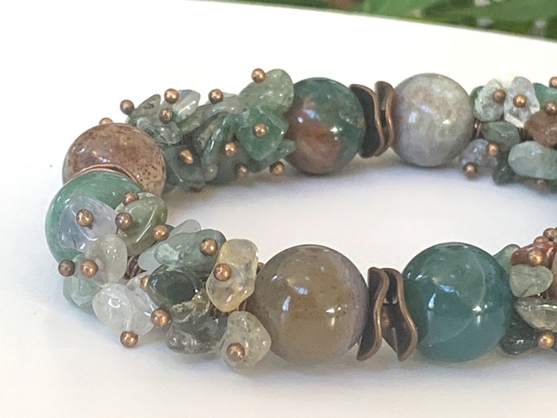 K&ouml;nnte beinhalten: Ein gr&uuml;nes, braunes und wei&szlig;es Edelstein-Armband mit Kupferdraht-Akzenten. Das Armband verf&uuml;gt &uuml;ber eine Vielzahl von Formen und Gr&ouml;&szlig;en von Edelsteinen, darunter runde, ovale und unregelm&auml;&szlig;ige Formen.
