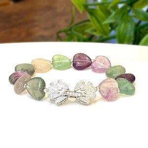 May include: A heart-shaped fluorite bracelet with a silver clasp. The bracelet features a variety of colors, including green, purple, and pink.
