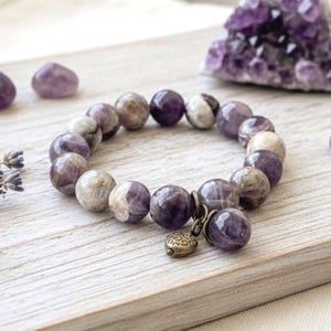 May include: A beaded bracelet featuring round, faceted amethyst stones in shades of purple and gray. The bracelet has a small, decorative charm and is displayed on a light-colored wooden surface. Amethyst crystals are visible in the background.