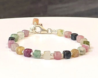 May include: A silver bracelet with square-shaped gemstones in various colors, including pink, green, yellow, and black.
