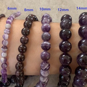 May include: A selection of gemstone bead bracelets, ranging from 4mm to 14mm in size. The bracelets are made of dark brown and purple beads. The sizes are indicated above each bracelet. The bracelets are shown on a person's arm.