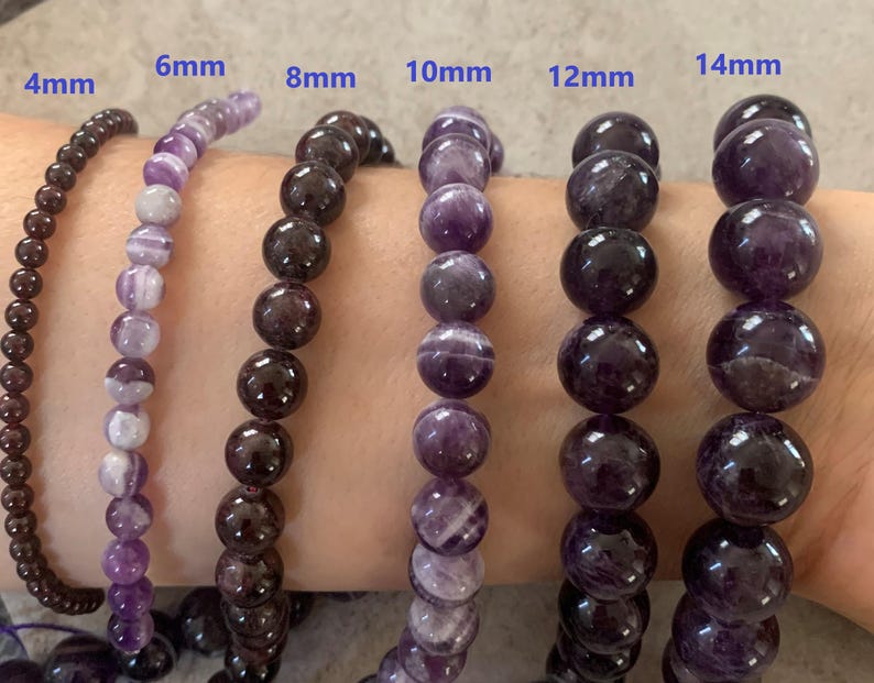 May include: A selection of gemstone bead bracelets, ranging from 4mm to 14mm in size. The bracelets are made of dark brown and purple beads. The sizes are indicated above each bracelet. The bracelets are shown on a person's arm.