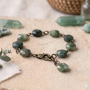 May include: A bracelet featuring faceted, sage green stone beads linked together with antique bronze-toned metal rings and a lobster claw clasp. A single matching stone bead hangs from the clasp, completing the circular design of the jewelry.