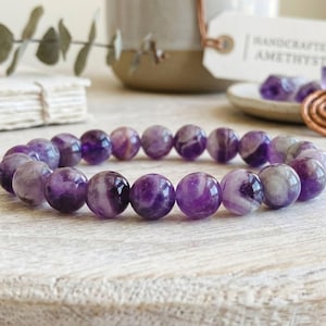 Puede incluir: Una pulsera el&aacute;stica compuesta por cuentas de amatista redondas y pulidas en varios tonos de morado intenso y blanco transl&uacute;cido. Las cuentas est&aacute;n ensartadas en un patr&oacute;n circular, mostrando variaciones de color naturales.