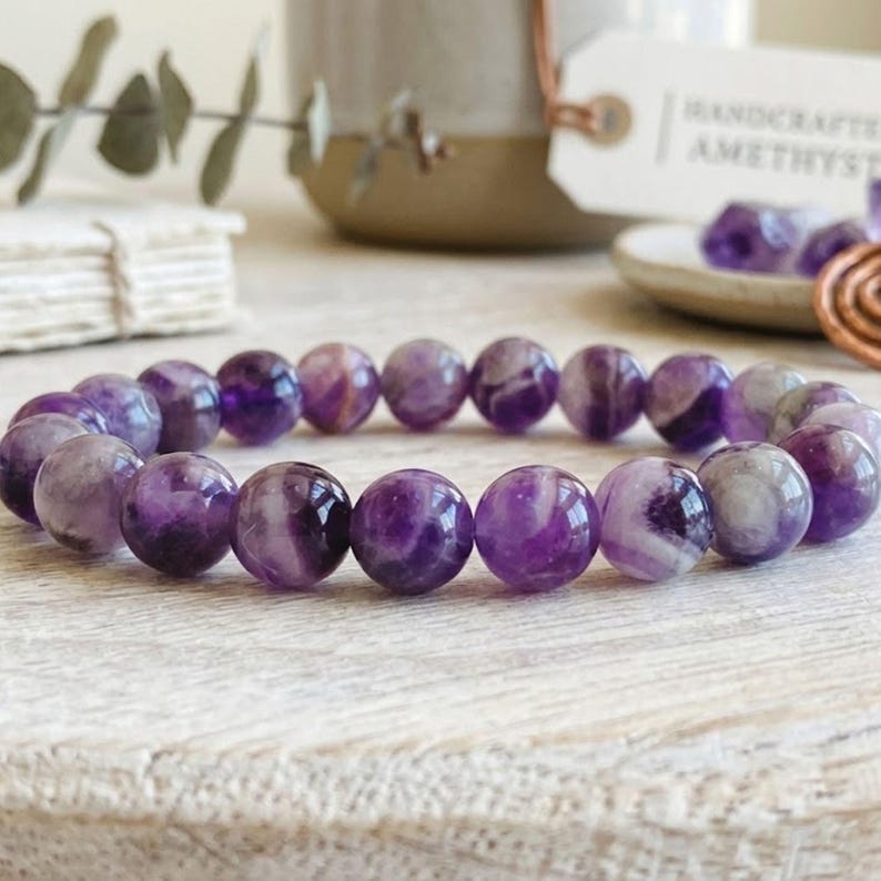 Puede incluir: Una pulsera el&aacute;stica compuesta por cuentas de amatista redondas y pulidas en varios tonos de morado intenso y blanco transl&uacute;cido. Las cuentas est&aacute;n ensartadas en un patr&oacute;n circular, mostrando variaciones de color naturales.