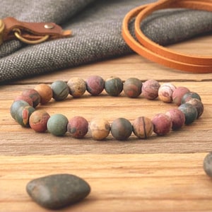 May include: A beaded bracelet composed of various earth-toned, round stones. The beads are primarily in shades of green, brown, and red, strung together to form a circular shape. The bracelet rests on a wooden surface, with leather accents in the background.