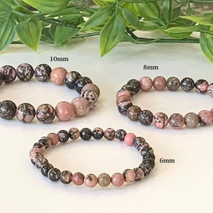 May include: Three beaded bracelets with pink, black, and grey stones. The bracelets are labelled with sizes: 10mm, 8mm, and 6mm. The background includes green leaves.