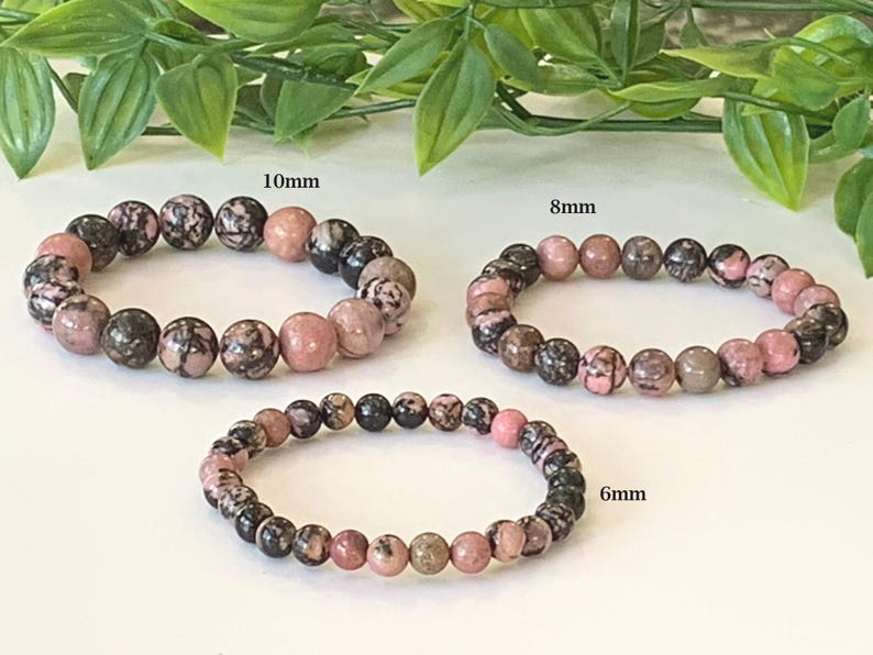 May include: Three beaded bracelets with pink, black, and grey stones. The bracelets are labelled with sizes: 10mm, 8mm, and 6mm. The background includes green leaves.