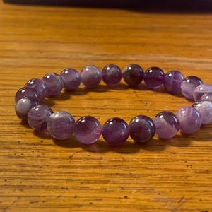Puede incluir: Una pulsera el&aacute;stica compuesta por cuentas de amatista redondas y pulidas en diversos tonos de morado y violeta. Las cuentas presentan variaciones de color naturales y cualidades transl&uacute;cidas, dispuestas en un bucle circular continuo.