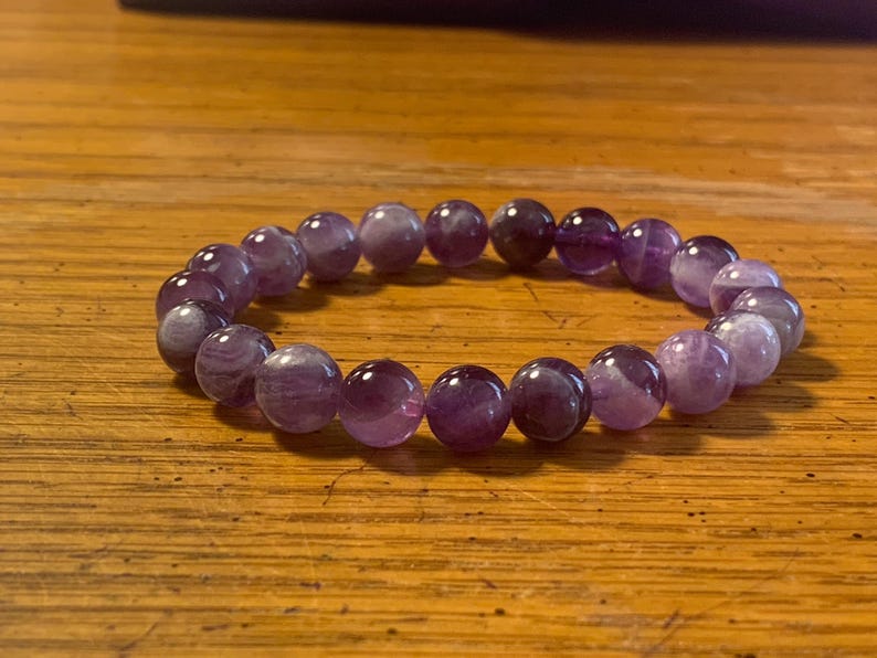 Puede incluir: Una pulsera el&aacute;stica compuesta por cuentas de amatista redondas y pulidas en diversos tonos de morado y violeta. Las cuentas presentan variaciones de color naturales y cualidades transl&uacute;cidas, dispuestas en un bucle circular continuo.