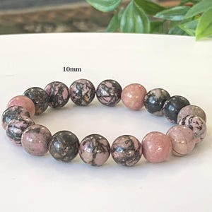 May include: A bracelet featuring 10mm round beads. The beads are a mix of pink and black, with a marbled pattern. The bracelet is presented on a white surface, with green foliage in the background.