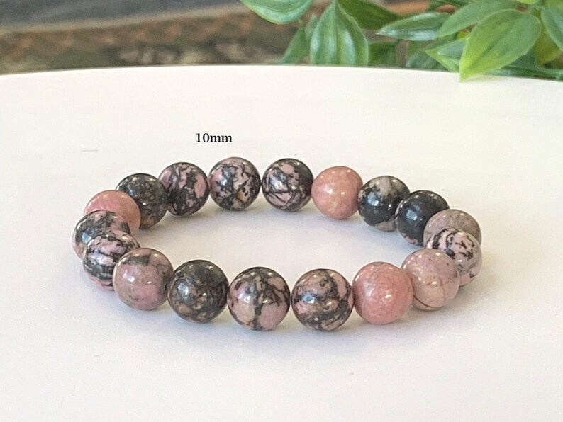 May include: A bracelet featuring 10mm round beads. The beads are a mix of pink and black, with a marbled pattern. The bracelet is presented on a white surface, with green foliage in the background.