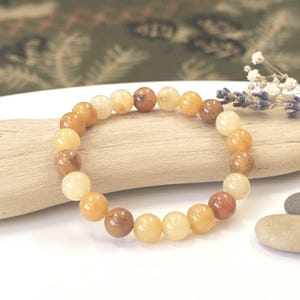 May include: A stretch bracelet composed of smooth, round beads in varying shades of amber, honey, and light brown. The beads exhibit a polished, semi-translucent finish, creating a warm, earthy color palette arranged in a circular pattern.