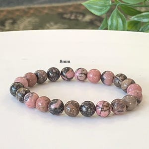 May include: A bracelet with 8mm beads in pink, grey, and black. The round beads are made of natural stone. The bracelet is displayed on a white surface.