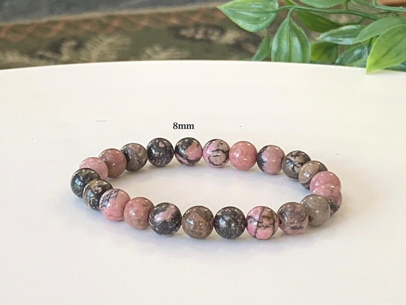 May include: A bracelet with 8mm beads in pink, grey, and black. The round beads are made of natural stone. The bracelet is displayed on a white surface.