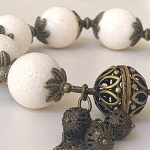Puede incluir: Pulsera de cuentas blancas con detalles en bronce. La pulsera presenta un gran dije de bronce adornado con un dise&ntilde;o de filigrana.