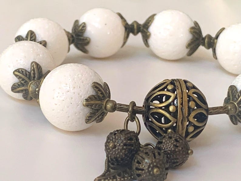 Puede incluir: Pulsera de cuentas blancas con detalles en bronce. La pulsera presenta un gran dije de bronce adornado con un dise&ntilde;o de filigrana.