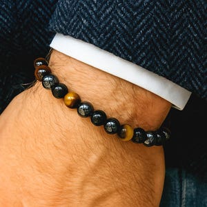 May include: A beaded bracelet featuring a mix of dark, glossy, and golden-brown spherical beads. The bracelet is worn on a wrist, with a dark blue blazer and white shirt cuff in the background. The beads appear to be approximately 0.4 inches in diameter.