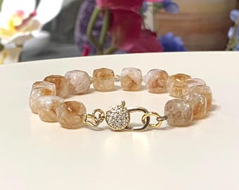 May include: A bracelet composed of faceted, translucent amber-colored cube beads with internal marbling. The bracelet features a gold-toned metal lobster claw clasp adorned with a circular pave setting of small, clear, sparkling crystals.