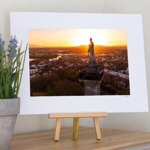 May include: A framed photograph of a cityscape at sunset, featuring a statue atop a tall structure. The sky is ablaze with orange and yellow hues. The photograph is displayed on a small wooden easel.