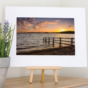 May include: A framed photograph of a sunset over a lake with a wooden fence in the foreground. The sky is a mix of pink, orange, and purple, and the water is calm and reflective. The fence is made of horizontal wooden planks and is partially submerged in the water.