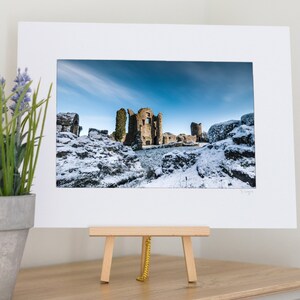 May include: A framed print of a snow-covered castle ruin under a bright blue sky. The print is displayed on a small wooden easel. A potted plant with purple flowers is to the left.