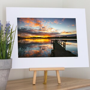 Puede incluir: Una fotografía enmarcada de un muelle de madera que se extiende hacia un lago al atardecer. El cielo es de un naranja y rosa vibrantes, reflejados en el agua. El muelle está hecho de madera desgastada y tiene una barandilla.
