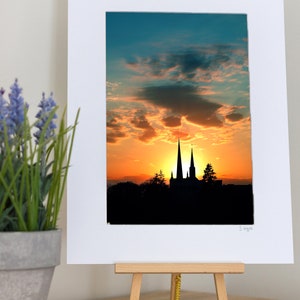 May include: A framed photograph of a church silhouetted against a vibrant sunset sky. The sky is a mix of orange, yellow, and blue hues with fluffy clouds. The church has two tall spires and is framed with a white mat.