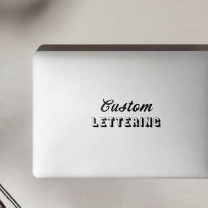 May include: A white laptop with a black sticker that says "Custom Lettering" on the lid. The laptop is on a white surface with a few brown acorns and a white piece of paper.