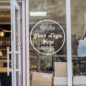 May include: A woman wearing a plaid shirt and grey apron stands outside a shop with a glass door. The door has a white circular sticker with the text "Your Logo Here" and floral designs. The shop window displays a variety of products, including bags, boxes, and jars.