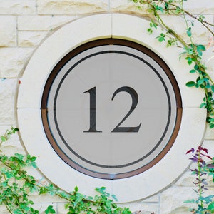 May include: A white circular sign with a black number 12 on a gray background. The sign is mounted on a brick wall with green vines growing around it.