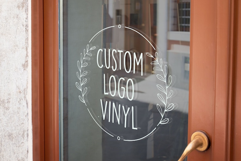 May include: A white vinyl sticker with a circle design and a wreath on either side. The text inside the circle reads "CUSTOM LOGO VINYL".