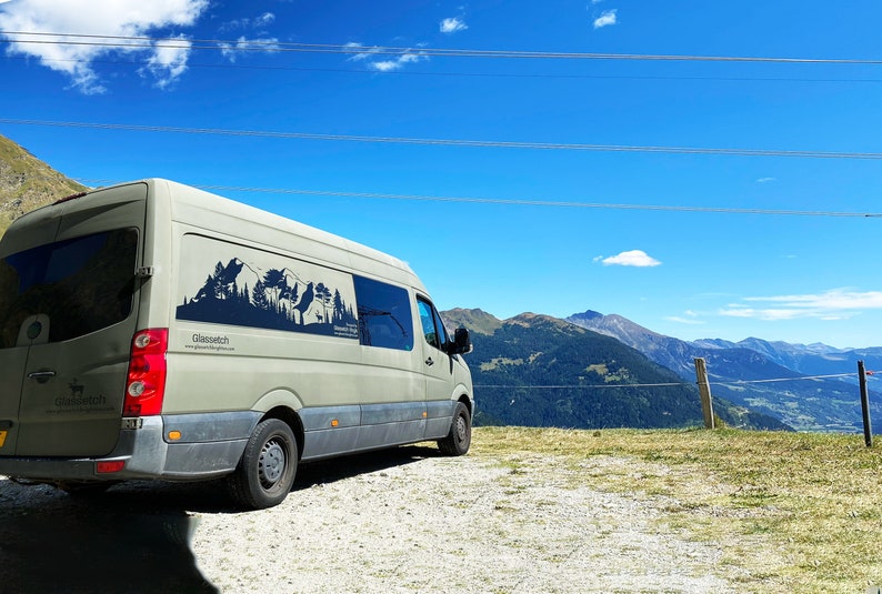 Op de afbeelding: Een witte bestelwagen met een zwarte berg silhouet afbeelding aan de zijkant. De bestelwagen staat geparkeerd op een onverharde weg met uitzicht op bergen op de achtergrond.