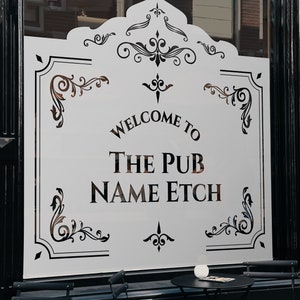 Op de afbeelding: Een wit bord met zwarte letters waarop staat "Welkom bij The Pub Name Etch". Het bord is versierd met sierlijke zwarte krulwerk en wordt getoond in een winkelruit.