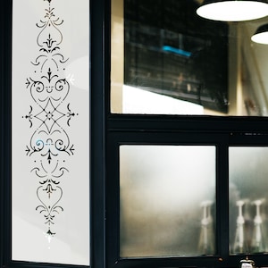 May include: A black framed window with a frosted glass pane featuring a decorative etched design. The window is divided into two sections, with the top section showing a blurry view of a room with a light fixture.