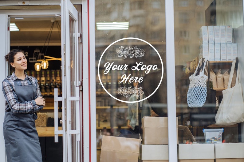 May include: A woman wearing a plaid shirt and grey apron stands outside a shop with a glass door. The door has a white circular sticker with the text "Your Logo Here" and floral designs. The shop window displays a variety of products, including bags, boxes, and jars.
