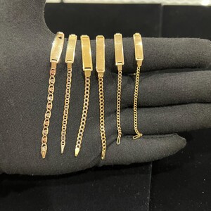 May include: Six gold-colored bracelets with rectangular nameplate-style clasps and various chain designs. The bracelets are displayed on a black surface, showcasing their polished finish and intricate chain patterns. The chains vary in style, including a mariner chain and a curb chain.