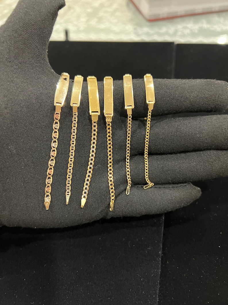 May include: Six gold-colored bracelets with rectangular nameplate-style clasps and various chain designs. The bracelets are displayed on a black surface, showcasing their polished finish and intricate chain patterns. The chains vary in style, including a mariner chain and a curb chain.