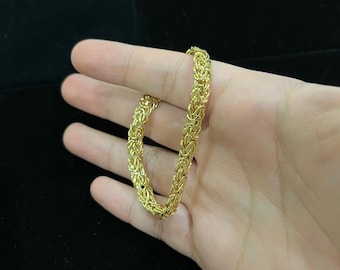 May include: A gold-coloured Byzantine chain bracelet. The bracelet features interlocking links, creating a textured and ornate design. The jewellery is held in a hand against a dark background.