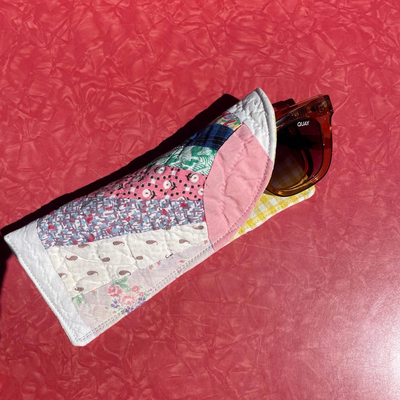 May include: A handmade quilted sunglasses case with a patchwork design in various colors and patterns, including pink, blue, and floral prints. The case holds a pair of brown sunglasses with the brand name "Quay" visible.