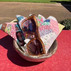 May include: A pair of brown-framed sunglasses rests atop a collection of colorful, patterned fabric pieces inside a ceramic bowl. The fabric pieces appear to be part of a quilt, featuring various floral and geometric designs in shades of pink, blue, and green.