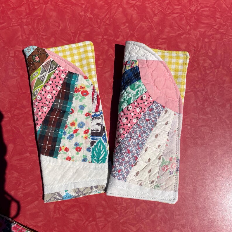 May include: Two handmade quilted fabric sleeves. Each sleeve features a patchwork design with various floral, plaid, and patterned fabrics in pink, blue, green, and white. The top edge is yellow gingham, and the bottom is white.