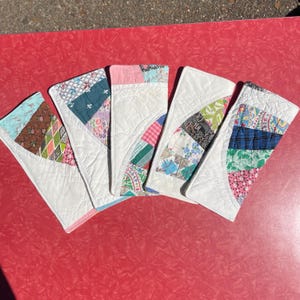 May include: Five handmade quilted coasters arranged in a fan shape. Each coaster features a white background with a colorful patchwork design in various patterns and colors, including pink, blue, green, and brown. The coasters are on a red surface.