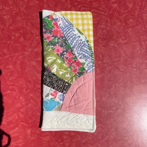 May include: A handmade, quilted wallet with a fan-shaped design. The wallet features a variety of fabric patterns, including floral, geometric, and gingham. The color palette includes pink, green, blue, and yellow. The wallet is rectangular and has a white border.