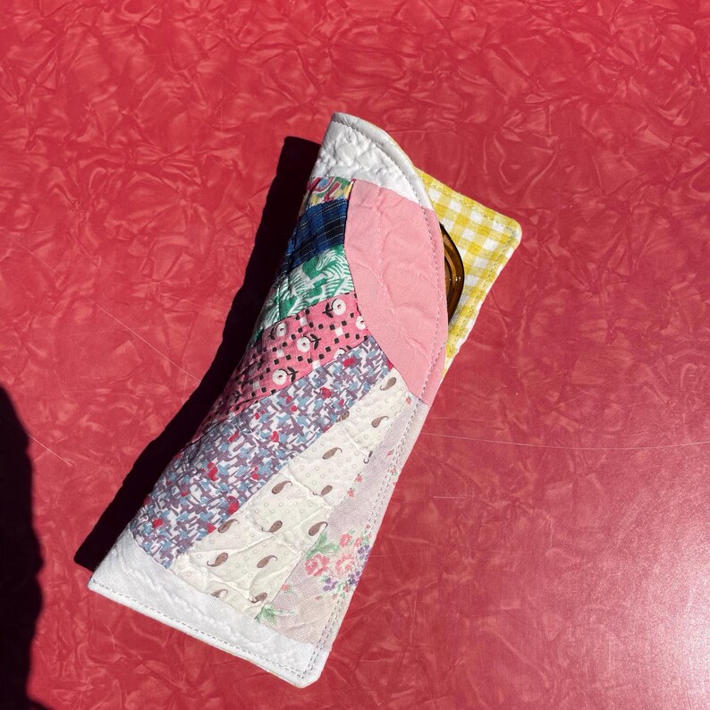 May include: A quilted glasses case with a patchwork design of various fabrics in pink, blue, green, and white. The case has a white border and a pink accent. A yellow and white checkered fabric is visible inside.