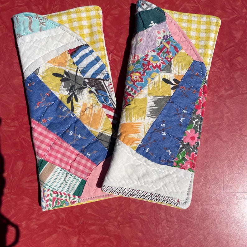 May include: Two quilted oven mitts with a patchwork design. The mitts feature a variety of fabrics in blue, yellow, pink, and white. The edges are trimmed with a yellow gingham fabric and white fabric. The mitts are designed for kitchen use.