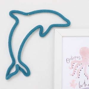 May include: A teal blue knitted dolphin wall hanging. The dolphin is in a jumping pose. The dolphin is hanging on a white wall next to a white picture frame with a pink octopus illustration.