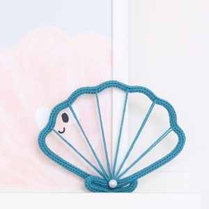 May include: A teal blue crocheted seashell with a white pearl in the centre. The seashell is a decorative item for a home or nursery.