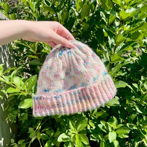 May include: A hand holding a pink, white and blue knitted beanie with a colourful pattern. The beanie has a folded brim.